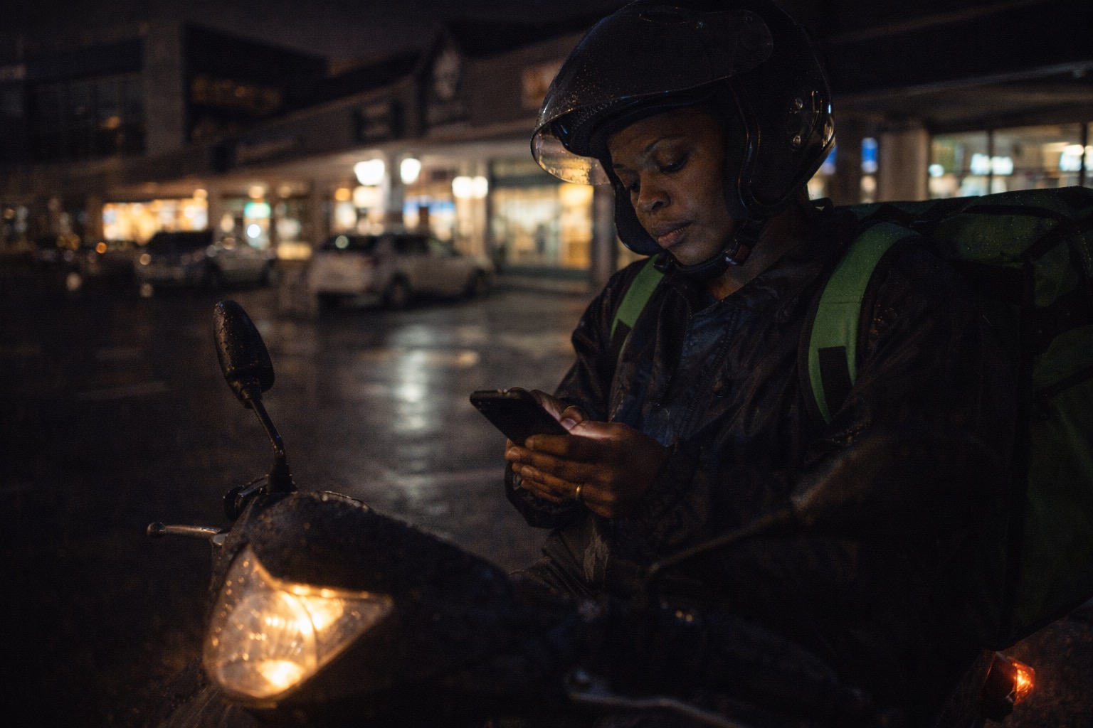A motorbike delivery rider in helmet on her bike at night, checking the app between drops